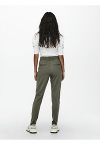 Femme aux longs cheveux bouclés, debout de dos, portant un haut blanc à manches bouffantes, un pantalon vert taille haute et des baskets blanches sur fond uni.