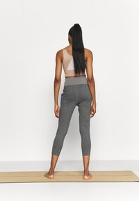 Mujer de pie descalza sobre una esterilla de yoga, de espaldas, vestida con un top corto beige y mallas gris ajustadas en una habitación blanca y sencilla.
