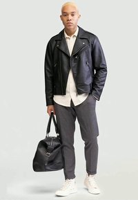 Black leather biker jacket, white shirt, gray patterned trousers, white sneakers, carrying a black duffel bag. Neutral background.