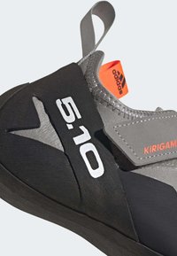 Sapatilha de escalada com um upper em preto e cinza, apresentando a marca "5.10" em branco e uma etiqueta laranja. Inclui uma superfície texturizada e uma alça.