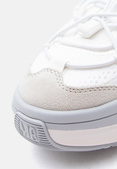 Λευκό και ανοιχτό γκρι αθλητικό παπούτσι σε κοντινή λήψη που δείχνει το μύτο από σουέντ, επάνω μέρος από δίχτυ, ελαστικά κορδόνια και λαστιχένια σόλα με το ανάγλυφο "MB" στην εμπρόσθια άκρη.