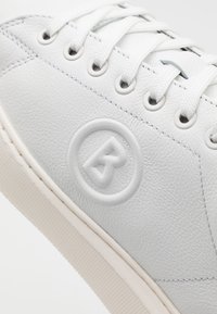 Białe skórzane sneakersy o teksturowanym wykończeniu, z płaskimi białymi sznurowadłami, okrągłym wytłoczonym logo i lekką gumową podeszwą.