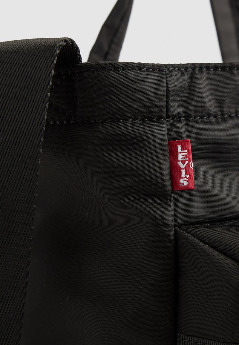 Bolso tote de tela negra con un diseño estructurado, que presenta una etiqueta del logo rojo de Levi's y detalles de costura prominentes. Asa texturizada.