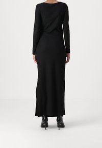 Schwarzes Langarm-Rippenkleid, figurbetonte Form, seitliche Schlitze am Saum und kontrastierendes glänzendes schwarzes High-Heel-Schuhwerk.