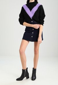 Schwarzer Strickpullover mit lila Chevron-Muster, kombiniert mit einem dunkelblauen Jeansrock mit Knopfleiste vorne und schwarzen Knöchelstiefeln mit Reißverschluss-Akzent.