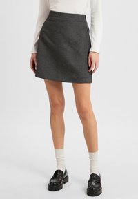 Grauer Woll-Minirock mit ausgestelltem Schnitt, mit seitlichem Knopfverschluss. Kombiniert mit schwarzen Slippern und weißen Knöchelsocken.