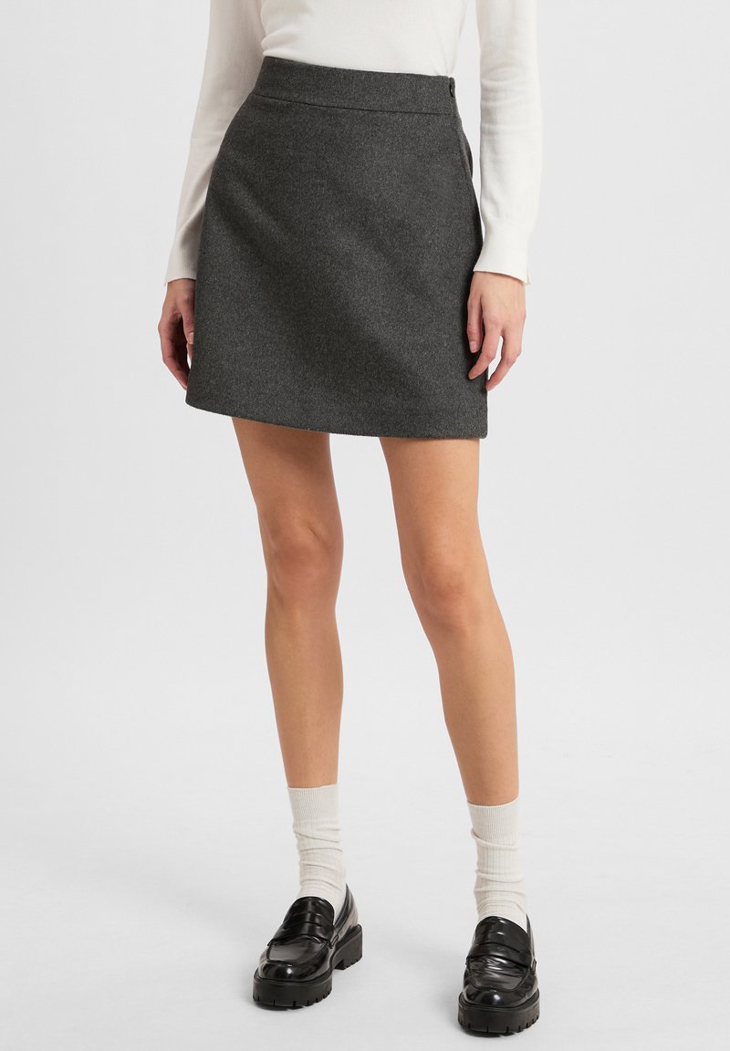 Grauer Woll-Minirock mit ausgestelltem Schnitt, mit seitlichem Knopfverschluss. Kombiniert mit schwarzen Slippern und weißen Knöchelsocken.