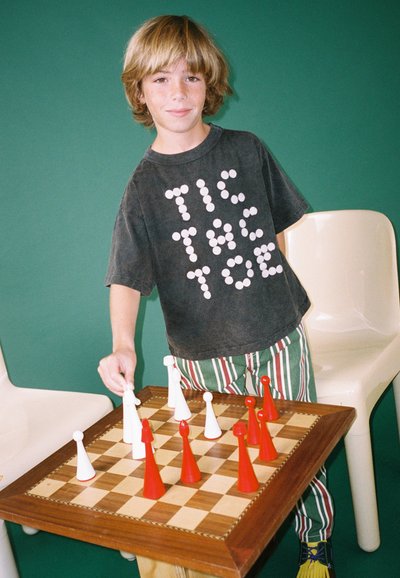 Schaakset met een houten bord en gepatternde stukken; speler in een grijs shirt met witte tekst, gestreepte broek en gele schoenen. Groene achtergrond.