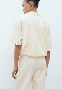 Light beige short-sleeve shirt with a loose fit and a collar. Smooth texture with subtle wrinkling; paired with matching light beige pants.