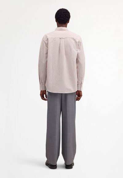 Mężczyzna stojący tyłem, ubrany w beżowo-białą pasiastą koszulę z długim rękawem, szare spodnie o szerokich nogawkach i czarne buty na białym tle.