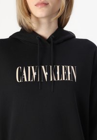 Črn pulover s kapuco, izdelan iz mehke tkanine, z izrazitim logotipom v svetlo bež barvi. Oblikovanje vključuje kapuco s vrvico in sproščen kroj.