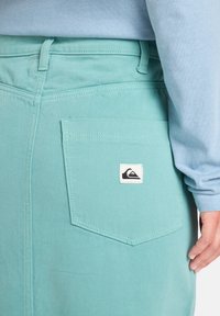Lichtblauw langemouwen shirt gelaagd over mintgroene denim met een achterzak voorzien van een zwart-witte logo-label.
