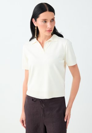 Femme aux longs cheveux foncés portant une chemise blanche à manches courtes avec col et un pantalon sombre, regardant légèrement vers sa gauche.
