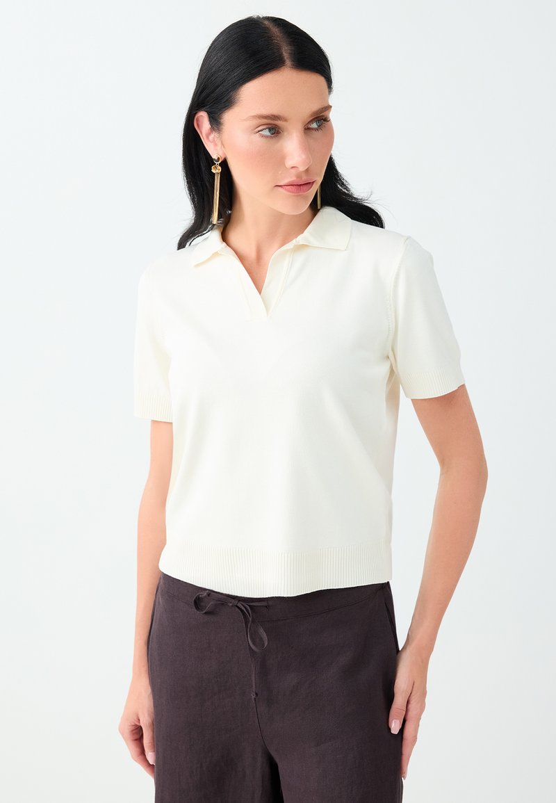 Femme aux longs cheveux foncés portant une chemise blanche à manches courtes avec col et un pantalon sombre, regardant légèrement vers sa gauche.