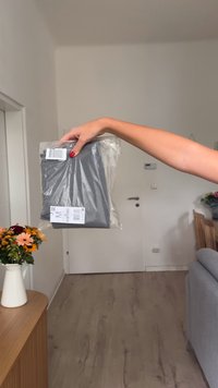 Hand med röd nagellack som håller ett förseglat grått klädförpackning med etiketter i en minimalistisk vardagsrumsmiljö.