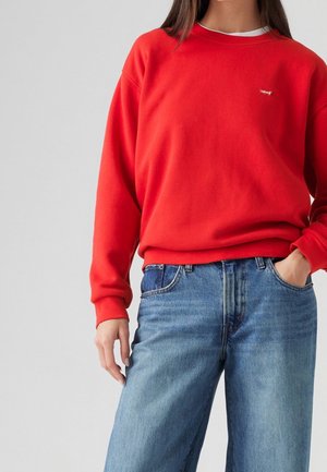 Personne portant un sweat-shirt Levi's rouge vif et un jean bleu avec une main dans la poche, sur un fond clair uni.