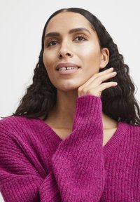 Pull en tricot fuchsia avec texture côtelée et encolure en V. Les manches longues tombent légèrement, mettant en valeur une apparence douce et confortable.