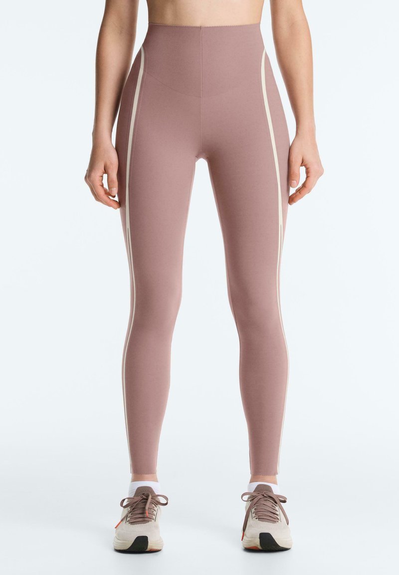 Leggings mauve taille haute avec des bandes latérales claires, portés avec des chaussettes blanches aux chevilles et des baskets beiges avec des lacets marron, montrés de la taille aux pieds.