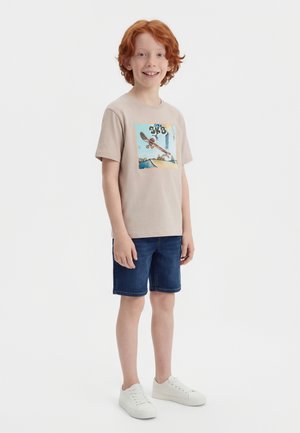 Uśmiechnięty rudowłosy chłopiec w beżowej koszulce z grafiką deskorolki, ciemnych dżinsowych szortach i białych trampkach stoi na tle białego tła.