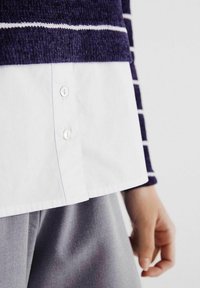 Námořnický pruhovaný pletený svetr přes bílou košili s knoflíky; viditelná šedá látka dole. Zaměření na knoflíky a kontrastní textury.