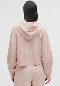 Hanorac scurt de culoare roz deschis, confecționat dintr-un material texturat, cu glugă, având detalii fine de cusătură și un logo mic pe spate.