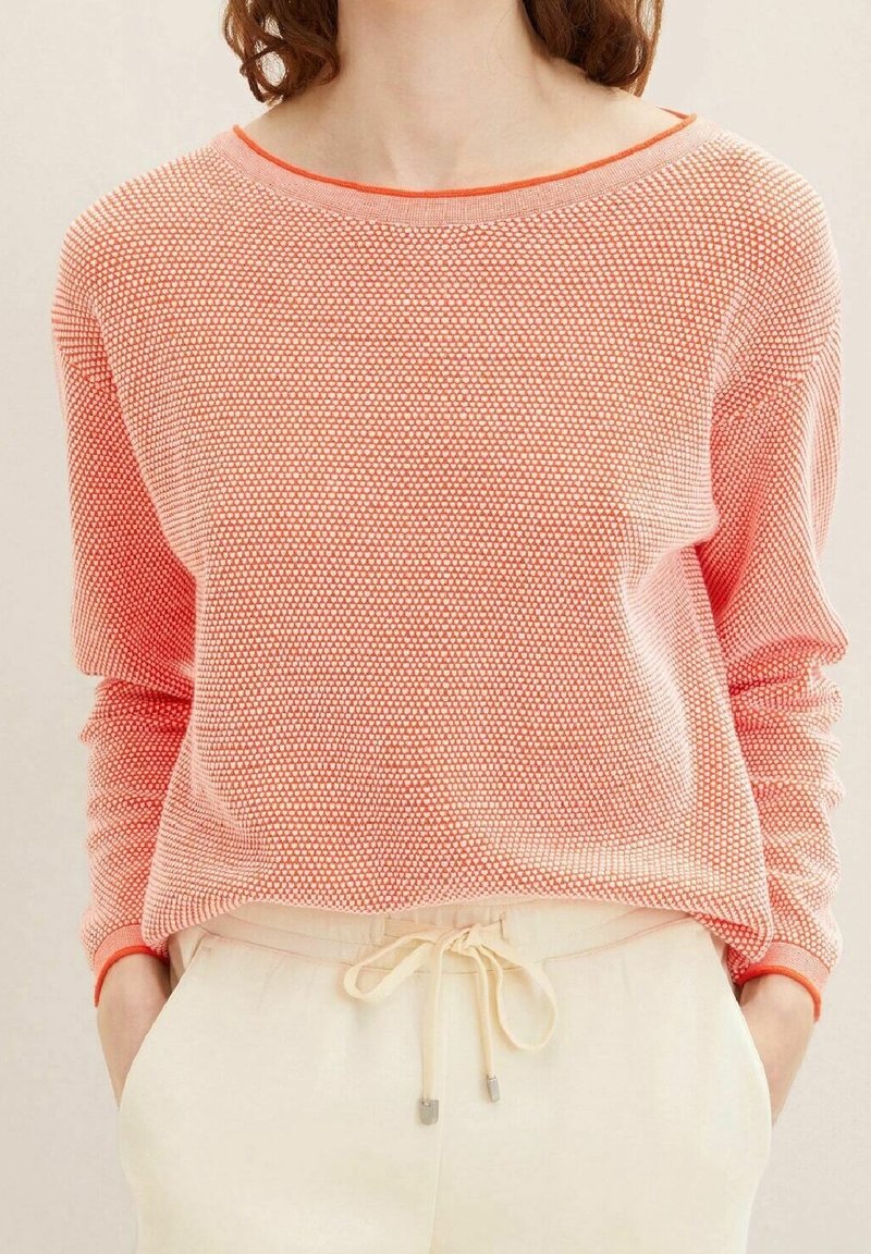 Femme portant un pull à manches longues à motifs corail, rentré dans un pantalon crème à cordon de serrage, les mains dans les poches, cadrée au niveau du cou et de la taille.