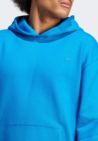 Person som bär en ljusblå huvtröja med bröstlogotyp, vänd något åt höger, med fokus på överkroppen och nacke.