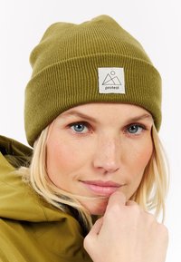 Woman with blue eyes wears olive green knit beanie with "protest" logo patch and matching jacket, resting chin on fist.