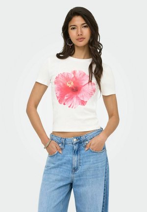 Mujer con cabello largo y oscuro que lleva un top corto blanco con un gran estampado de flores rosas y unos vaqueros de tiro alto de color azul claro, manos en los bolsillos.