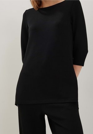 Femme portant un haut noir uni à manches longues et un pantalon noir assorti, debout devant un fond neutre.