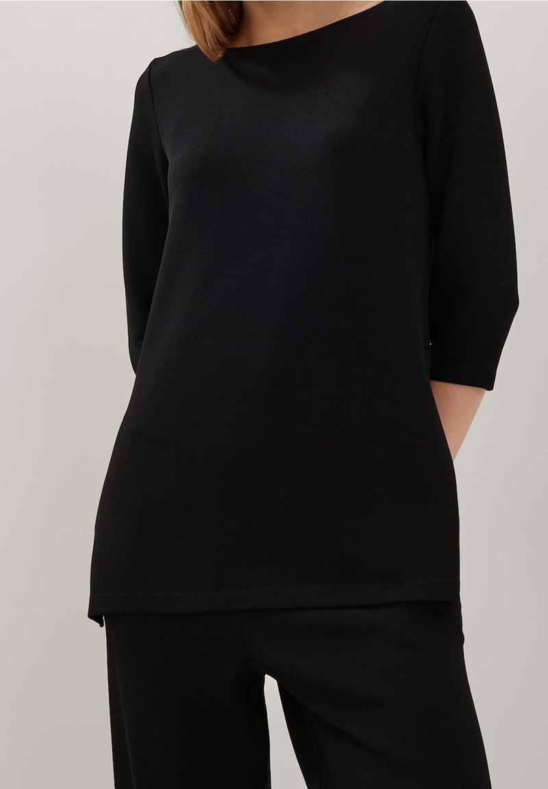 Femme portant un haut noir uni à manches longues et un pantalon noir assorti, debout devant un fond neutre.