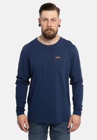 Langärmliges, navy-blaues T-Shirt aus Baumwolle mit einer Brusttasche und einem Etikett. Glatte Textur und entspannte Passform, kombiniert mit blauen Jeans.