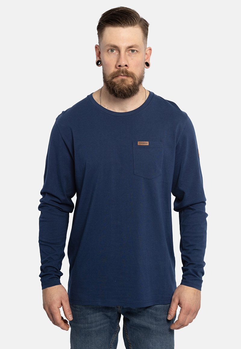 Langärmliges, navy-blaues T-Shirt aus Baumwolle mit einer Brusttasche und einem Etikett. Glatte Textur und entspannte Passform, kombiniert mit blauen Jeans.