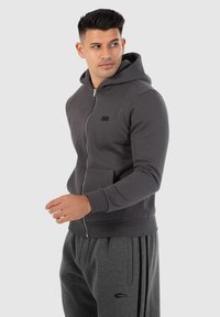 Sweat à capuche zippé gris avec une poche frontale, des poignets côtelés et une capuche. Porté avec un pantalon de jogging gris assorti avec des rayures noires.