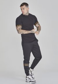Zwarte sport t-shirt met oranje logo op de mouwen, gecombineerd met zwarte joggers. Kenmerkende tattoo's en sneakers met witte accenten.
