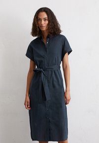 Abito camicia in lino blu scuro con maniche corte, chiusura con bottoni sul davanti e cintura da annodare in vita; presenta un taglio diritto lungo fino alla caviglia.