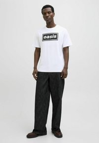 Balta marškinėliai su juodu "oasis" logotipu, derinami su laisvos formos tamsiai pilkais raštuotais kelnais ir tamsiai batais. Paprastas ir neformalus dizainas.