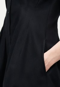 Χέρι μερικώς εισαχθέν στην πλαϊνή τσέπη μαύρου ραμμένου ενδύματος με ραφές σε λείο ύφασμα.