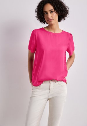 Femme aux cheveux bouclés portant un chemisier rose vif à manches courtes et un pantalon blanc, debout devant un fond clair uni.