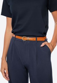 Cintura in pelle tan con fibbia in ottone, indossata su pantaloni navy e una t-shirt nera. La cintura presenta perforazioni visibili e dettagli di cucitura.