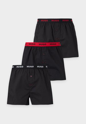 Muž bez košele stojí, má oblečené čierne boxerky s červeným logom "HUGO" na páse, pozerá doľava pred jednofarebným pozadím.