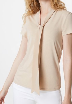 Beige Kurzarmoberteil mit V-Ausschnitt und einem langen Bindeband in der Mitte. Glatte Textur und figurbetonter Schnitt. Kombiniert mit weißen Hosen.