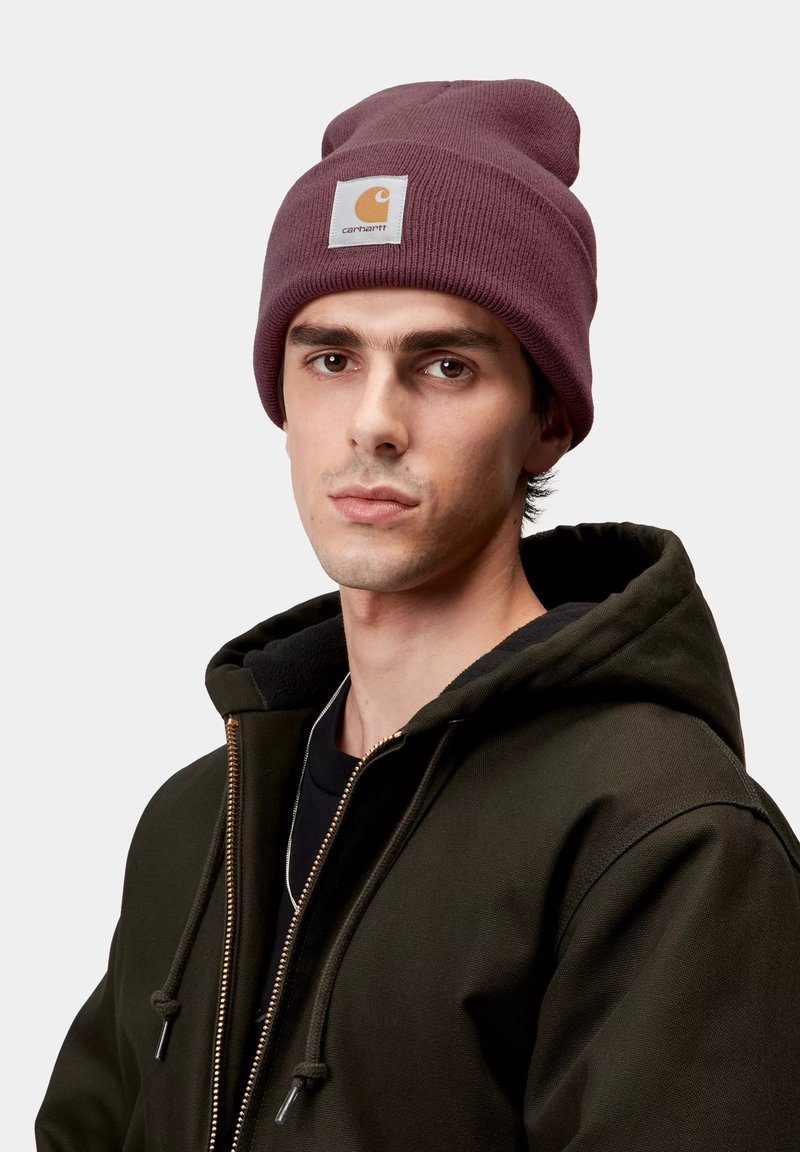 Giovane uomo che indossa un berretto Carhartt viola e una felpa con zip verde scuro con cappuccio, che guarda dritto davanti a sé su uno sfondo bianco.