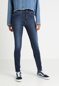 Vaqueros ajustados de mezclilla oscura con una altura media, combinados con una chaqueta de mezclilla recortada de color azul claro. El calzado presenta zapatillas oscuras con acentos blancos.