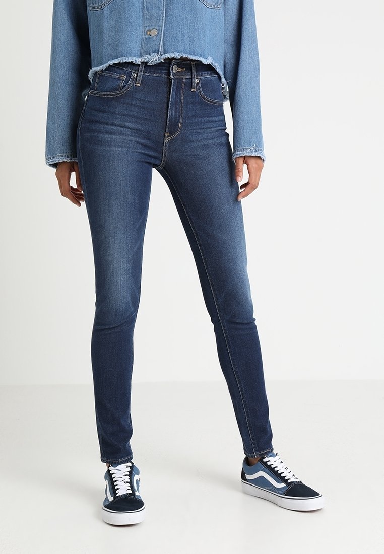 Vaqueros ajustados de mezclilla oscura con una altura media, combinados con una chaqueta de mezclilla recortada de color azul claro. El calzado presenta zapatillas oscuras con acentos blancos.