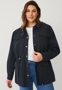 Giacca nera morbida con due tasche sul petto, bottoni a pressione e vita regolabile con cordino. Abbinata a una camicia bianca e jeans blu.