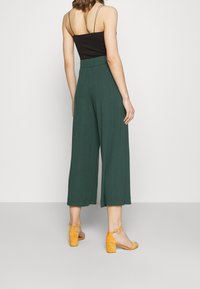 Nainen seisoo selkä otettuna, päällään tummanvihreät leveälahkeiset housut, musta hihaton toppi ja keltaiset nilkkaremmiset blokkikorkokengät.