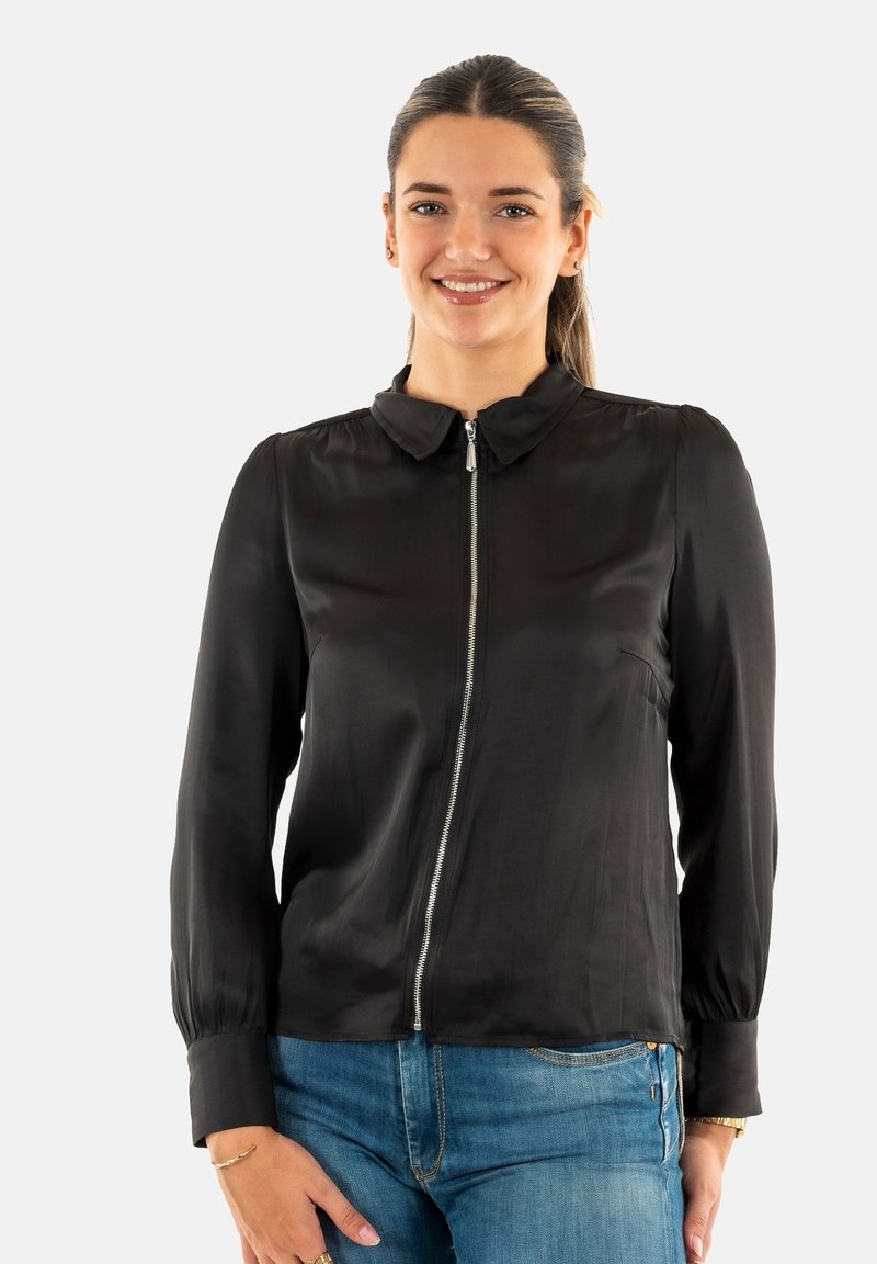 Blouse zippée noire avec un col rond, manches longues et poignets plissés ; fabriquée en tissu lisse et glossy ; portée avec un jean bleu.