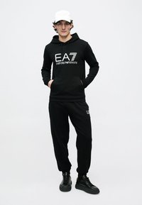 Fekete kapucnis pulóver az "EA7 Emporio Armani" logóval, kenguruzsebbel, fekete melegítőnadrággal és fekete tornacipővel párosítva. Egyszerű dizájn és egységes szín.