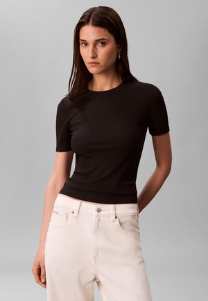 Kobieta ubrana w dopasowaną czarną bluzkę z krótkim rękawem i wysokim stanem jasnobeżowe spodnie, stojąca na jednolitym szarym tle.